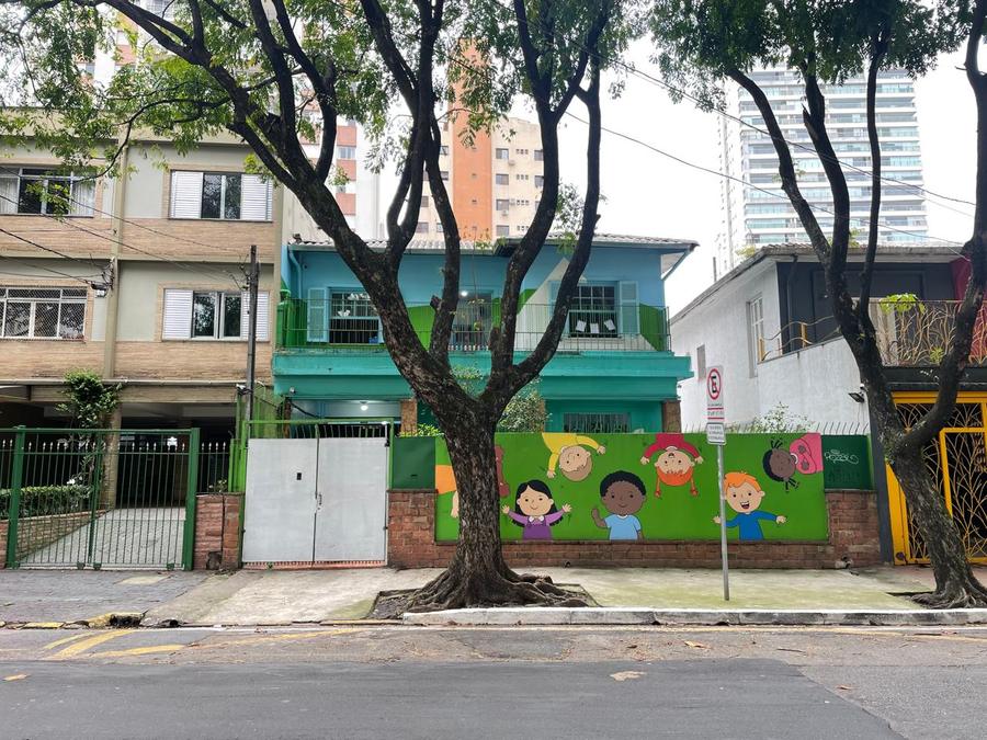 Fachada da Escola Quintal da Vovó na Rua Padre Carvalho, vista da outra calçada: o sobrado azul e verde, o muro com as crianças pintadas e duas árvores na frente.