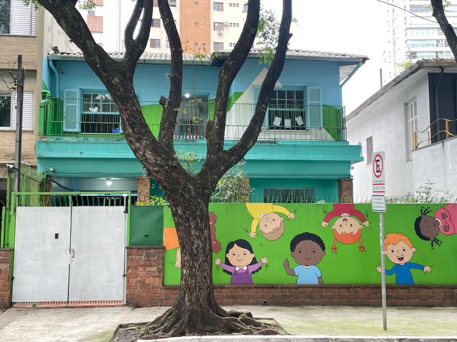 A mesma fachada mais de perto: o muro verde com o desenho das crianças, o portão de entrada e a árvore em frente à porta.